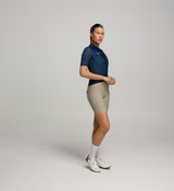 Woman wearing a navy cycling jersey and beige riding bib shorts on a gray background